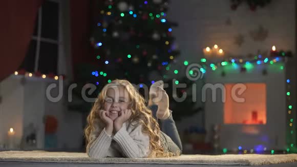 女孩躺在地板上靠近闪闪发光的圣诞树和舒适的壁炉庆祝活动视频的预览图