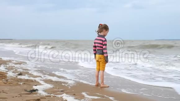 滑稽的女孩在沙特和黄裤子看着波浪在海洋站在水附近海上的弱风视频的预览图