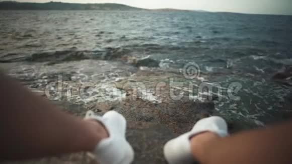 在海浪的背景下穿着白色运动鞋的腿在岩石海滩上第一人称射击焦点移到视频的预览图