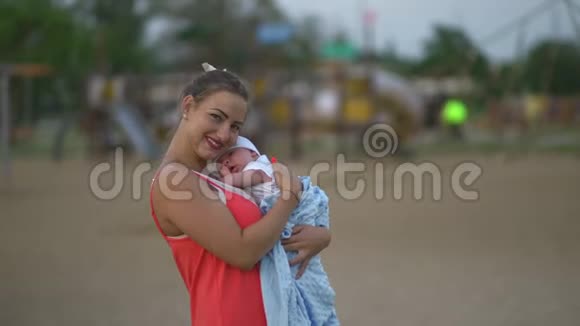 年轻的母亲抱着她的孩子在城市公园玩耍她站在那里穿着鲜红的裙子儿子穿着视频的预览图