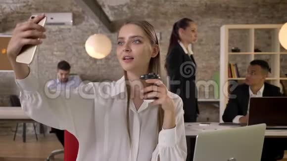 年轻漂亮的女商人正在办公室的智能手机上用咖啡做自拍她的同事们正在与视频的预览图