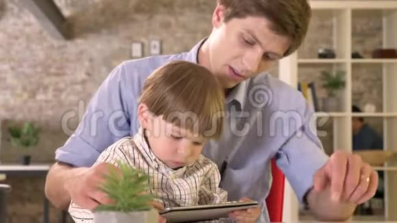 年轻的父亲和他的小儿子一起看平板电脑孩子被他的父亲惹恼了坐在现代的砖制办公室里视频的预览图