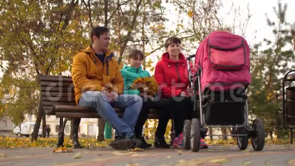 已婚夫妇带着孩子坐在秋天公园的长凳上带着婴儿车和谈话视频的预览图