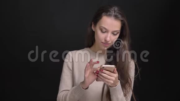 年轻的黑发白种人长发女孩在黑色背景下专心观看智能手机的肖像视频的预览图