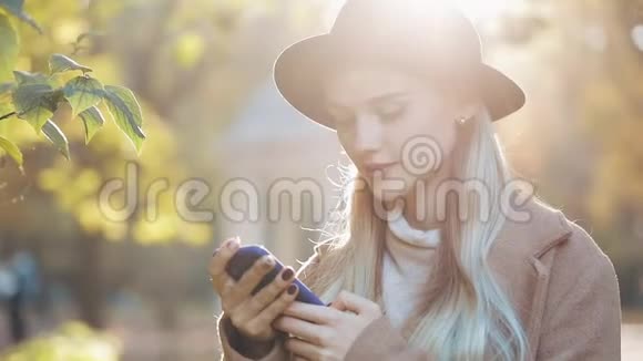 年轻女子穿着外套用智能手机站在秋天公园女孩看着镜头户外技术视频的预览图