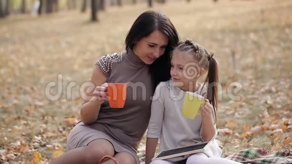 快乐的母女正在公园里坐在格子布上喝茶一家人在城市公园野餐视频的预览图