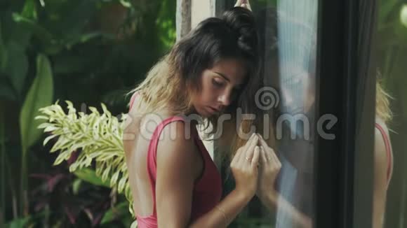 一个穿比基尼的模特站在她舒适的热带房子的玻璃门附近悲伤地低头抬起她的目光视频的预览图