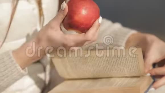 美丽的年轻女孩在公园里的一棵树下吃一个红色的苹果在水边看书双手合拢视频的预览图