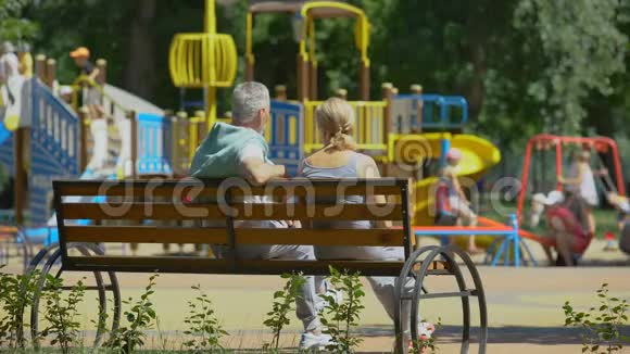 老两口坐在操场旁的长凳上看着孙辈们玩耍视频的预览图