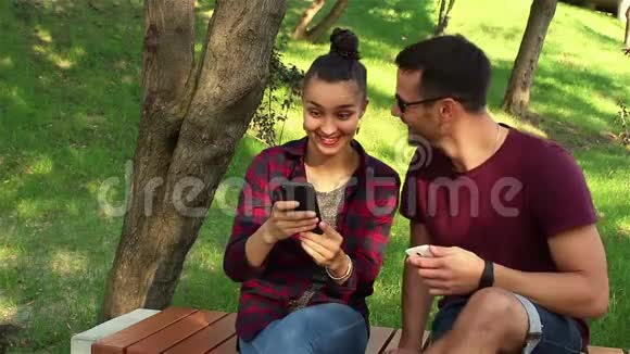 恋爱中的情侣坐在公园的长凳上在智能手机上互相展示照片视频的预览图