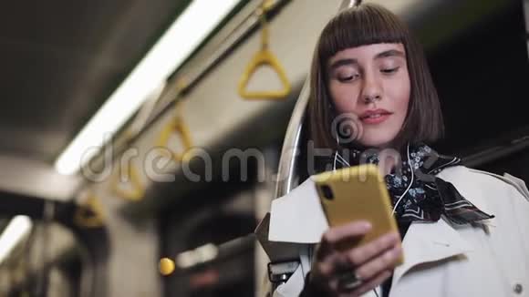 戴着耳机乘坐公共交通工具听音乐和浏览黄色智能手机的微笑年轻女性的肖像视频的预览图