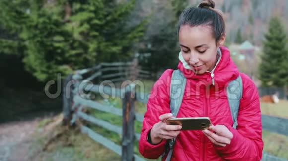 穿着红色夹克和小背包的漂亮女徒步旅行者正在用电话站在树林背景上视频的预览图