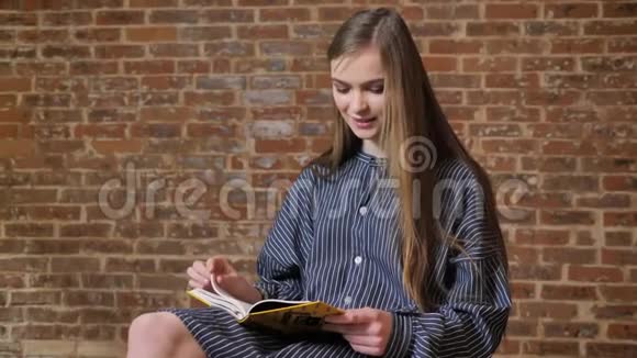 年轻漂亮的女孩坐着看书看着相机微笑着放松的概念砖背景视频的预览图