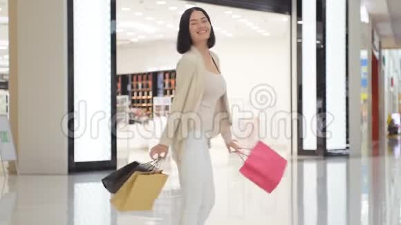 女人穿着柔软的彩色牛仔裤和夹克双手拿着购物袋在商场里走来走去视频的预览图
