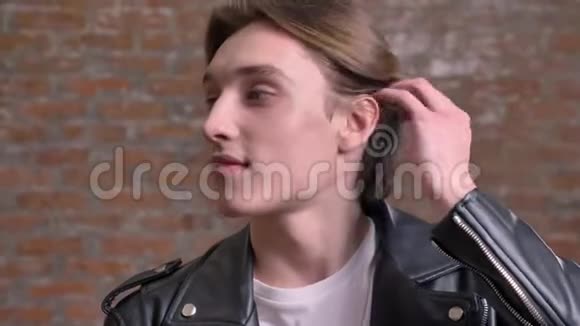 年轻的帅哥穿着黑色皮夹克摸着他的头发对着镜头微笑被孤立在砖背景上视频的预览图