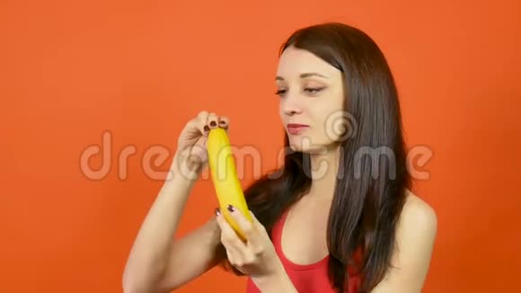 美丽的布鲁内特年轻女子手里拿着黄色香蕉然后吃它在明亮的橙色背景热带视频的预览图