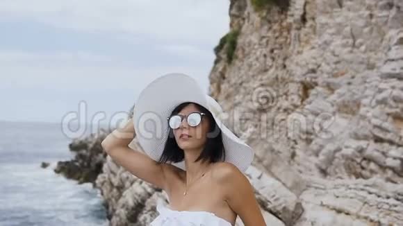 美丽的年轻女子穿着比基尼戴着大帽子和太阳镜在海滩上放松在海边的岩石背景下放松巴德瓦视频的预览图