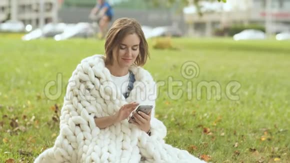 一个裹着美利奴格子的小女孩在一个城市公园的草地上打电话留言视频的预览图