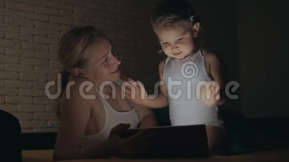 晚上一个小女孩在她母亲旁边的床上挥舞着手臂看着电子平板电脑视频的预览图