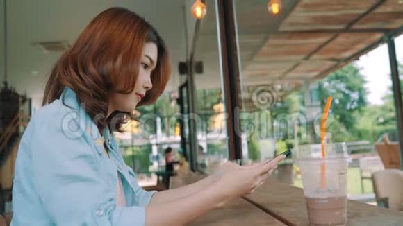 快乐的亚洲年轻女人坐在咖啡馆里喝着冰绿茶用智能手机聊天阅读和发短信视频的预览图