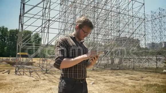 戴着眼镜的年轻人用平板电脑看着背景上的金属建筑站在稻草场上严肃而严肃视频的预览图