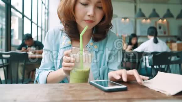 快乐的亚洲年轻女人坐在咖啡馆里喝着冰绿茶用智能手机聊天阅读和发短信视频的预览图