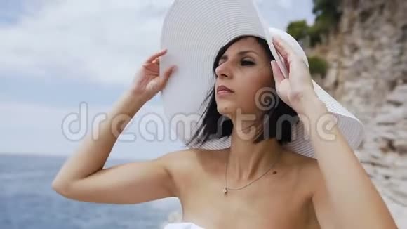 一位身穿白色泳衣和帽子的美丽年轻女子的肖像背景是海蓝色的水和层状岩石概念视频的预览图