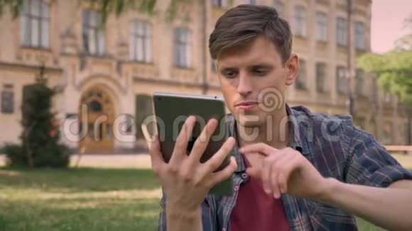 年轻的帅哥坐在公园的草地上轻敲平板电脑看蓝屏看镜头放松概念视频的预览图