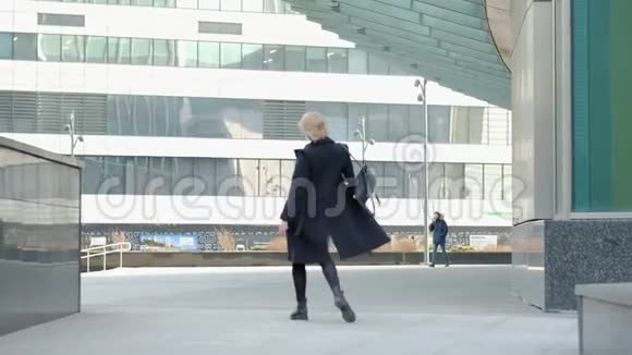 带背包行走的金发女人戴着黑帽子对着建筑玻璃墙后侧视慢动作视频的预览图