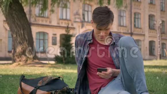 年轻的帅哥坐在公园的草地上看智能手机上的互联网页面看相机放松一下视频的预览图