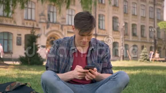 年轻的帅哥坐在公园里的草地上敲着智能手机对着镜头狂吠放松概念交流视频的预览图
