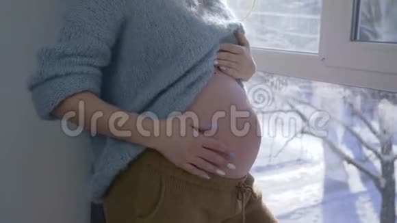 怀孕的夫妇爱抚肚子爸爸妈妈站在大窗户旁双手放在赤裸的大肚子上视频的预览图