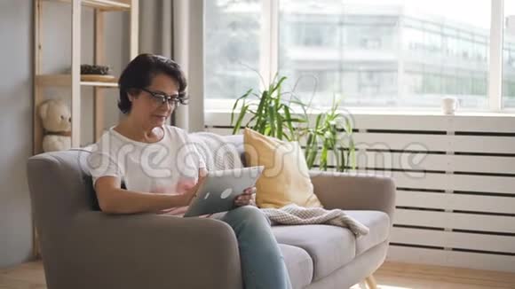 中年妇女浏览网页使用平板电脑坐在公寓的沙发上视频的预览图