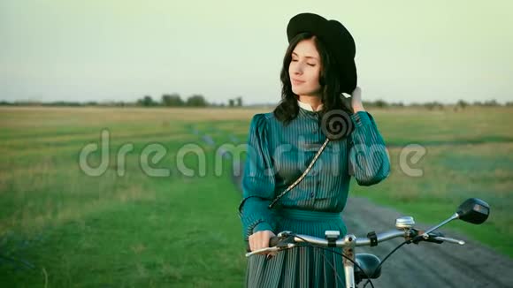 草原上一个穿着老式连衣裙戴帽子骑着自行车的黑发女人的肖像视频的预览图