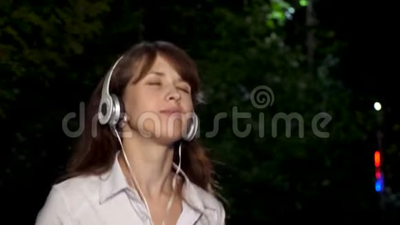 美丽的女孩戴着耳机长发唱歌跳舞微笑着晚上在公园享受音乐特写视频的预览图