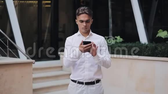 年轻成功商人的肖像智能手机站在办公楼附近看着摄像机视频的预览图