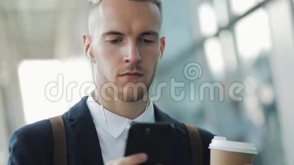 年轻的商人站在办公楼附近用带耳机的智能手机和喝咖啡商人视频的预览图