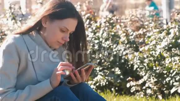 年轻漂亮的黑发女人坐在春天公园的草地上用手机看社交媒体视频的预览图