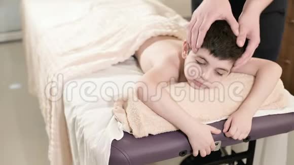 一位男性理疗师按摩师为躺在按摩床上的一个微笑的小男孩做一个疗愈放松的按摩头视频的预览图