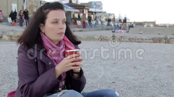 4一位穿春衣的漂亮女士端着一杯热饮料坐在海滩上视频的预览图
