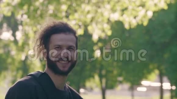 一个留胡子的年轻人站在阳光明媚的夏天公园里微笑着然后回头4K半转侧视频的预览图