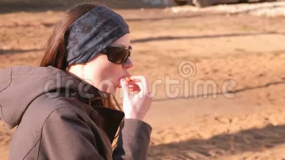 年轻的黑发女人戴着太阳镜在公园的长凳上吃橘子侧视视频的预览图