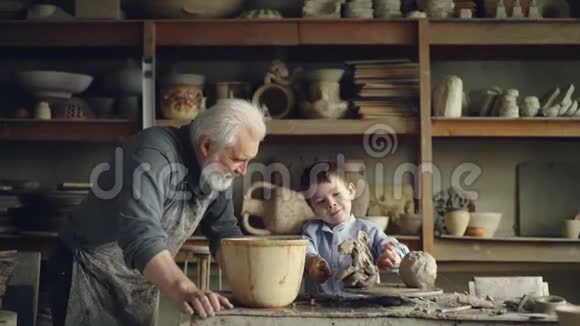 可爱的小男孩正在他祖父的工作室里玩粘土制作陶瓷玩具白发苍苍的胡须男视频的预览图