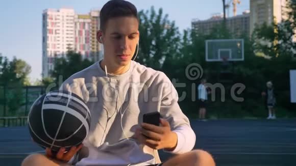 一个英俊的年轻人坐在篮球场上打电话看着摄像机拿着球人们在玩视频的预览图