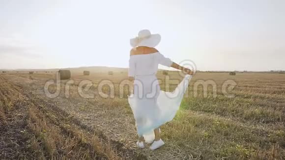 迷人的女孩穿着一件长裙子戴着围巾的大帽子在日落的夏天在田野里旋转年轻美女视频的预览图