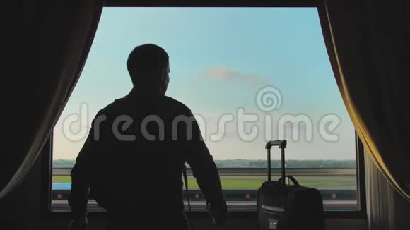 一个年轻人看着从旅馆房间窗户起飞的飞机带着一个手提箱离开登机视频的预览图
