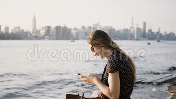 迷人的白种人女孩戴太阳镜使用智能手机信使应用程序环顾纽约河滩4K视频的预览图