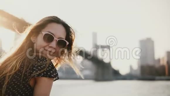 快乐的欧洲女孩戴着太阳镜微笑在纽约布鲁克林大桥享受令人惊叹的河流日落看着相机视频的预览图
