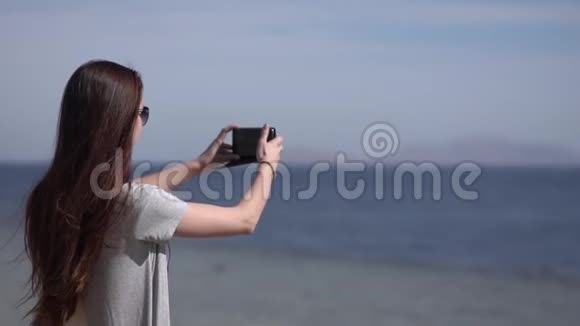 年轻的女孩在她的智能手机上拍摄大海的照片视频的预览图