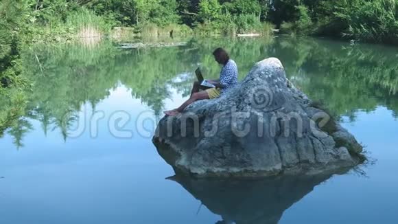 一个年轻的自由职业者坐在湖中央的石头上手提电脑自由职业概念出差工作无线无线视频的预览图
